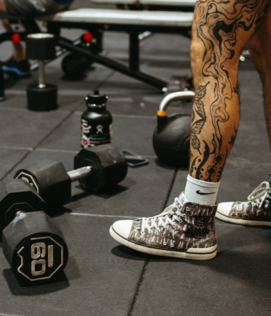 A person with tattooed legs and patterned sneakers stands near a 60-pound dumbbell, a kettlebell, and a water bottle in a gym setting, ready for their next climbing-inspired workout.