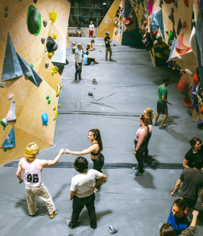 Indoor climbing gym with several people climbing or standing on padded floors. Two women in the center are fist-bumping, while others are talking, observing, or preparing to climb the colorful climbing walls.