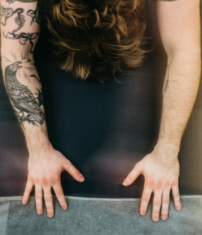 A person with tattoos on their arms lies face down on a yoga mat, hands flat on the ground as if preparing for climbing, brown hair spread out. The image is viewed from above.