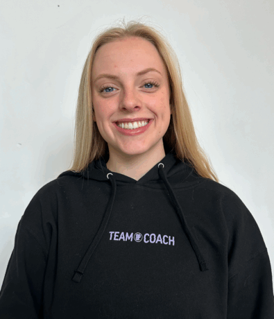 A young woman with long blonde hair smiles at the camera. She is wearing a black hoodie that says TEAM COACH in white letters, representing the Climb Team in Minneapolis, and stands against a plain white background.