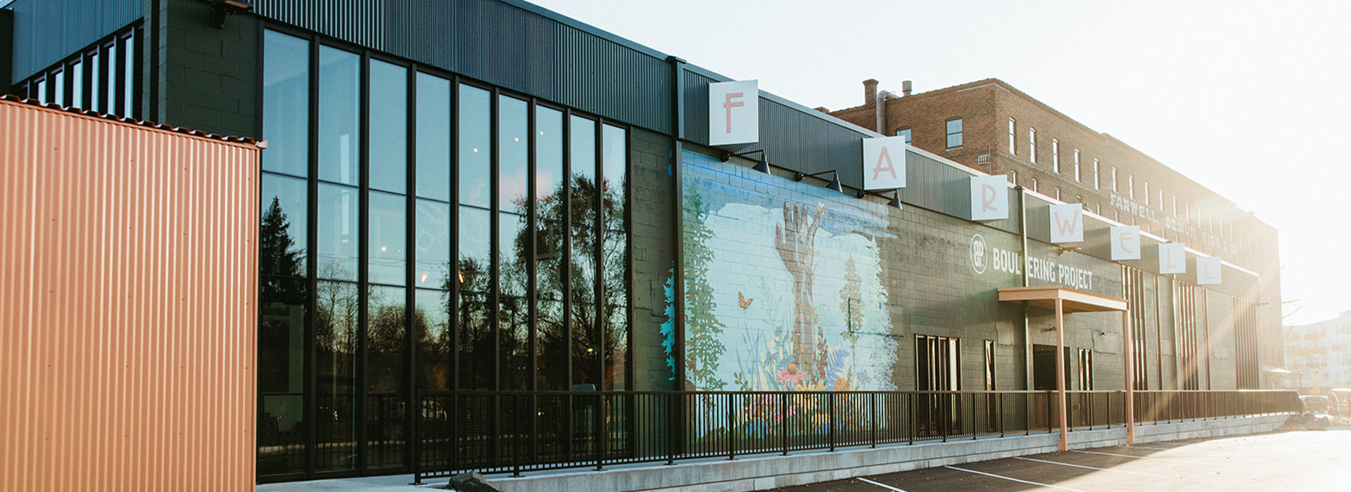 A modern building with large glass windows, a colorful mural of hands and flowers, and white letters above spelling FARWELL. The sun is shining on the empty parking lot in front of this former St Paul rock climbing gym.