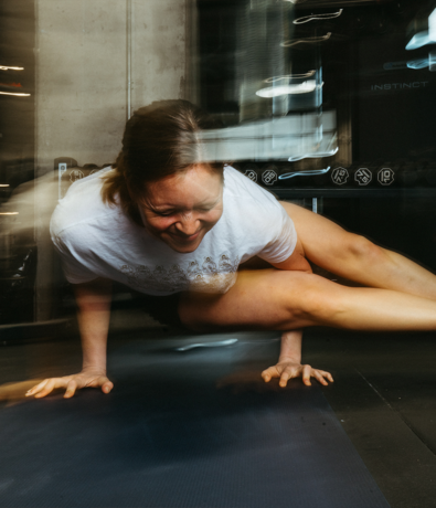 A woman balances on her hands in a yoga pose on a mat at a gym, smiling with focus. Motion blur is visible, suggesting energy and movement in the scene.