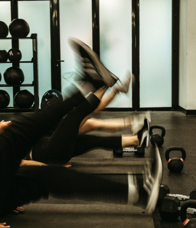 Three people exercise indoors, lying on mats and performing leg raises in a gym. Medicine balls and kettlebells are visible in the background. The image captures motion blur in their legs.
