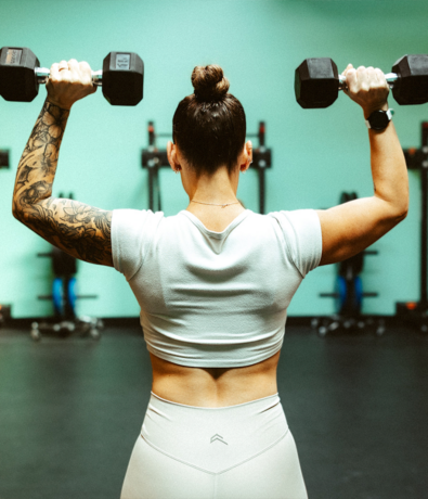 A woman with a tattooed arm and a bun hairstyle, wearing a white crop top and leggings, lifts two dumbbells above her shoulders in an Austin climbing gym. The background features gym equipment and a teal wall.