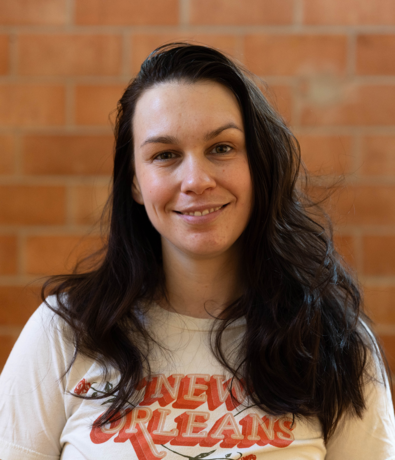 A woman with long dark hair smiles softly at the camera while wearing a white New Orleans T-shirt. The background, reminiscent of an Austin climbing gym, consists of an orange brick wall.