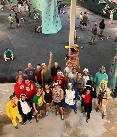 A group of people in colorful costumes pose for a photo at an indoor rock climbing gym, celebrating Community & Events at Austin Bouldering Project, with more climbers and large climbing walls visible in the background.