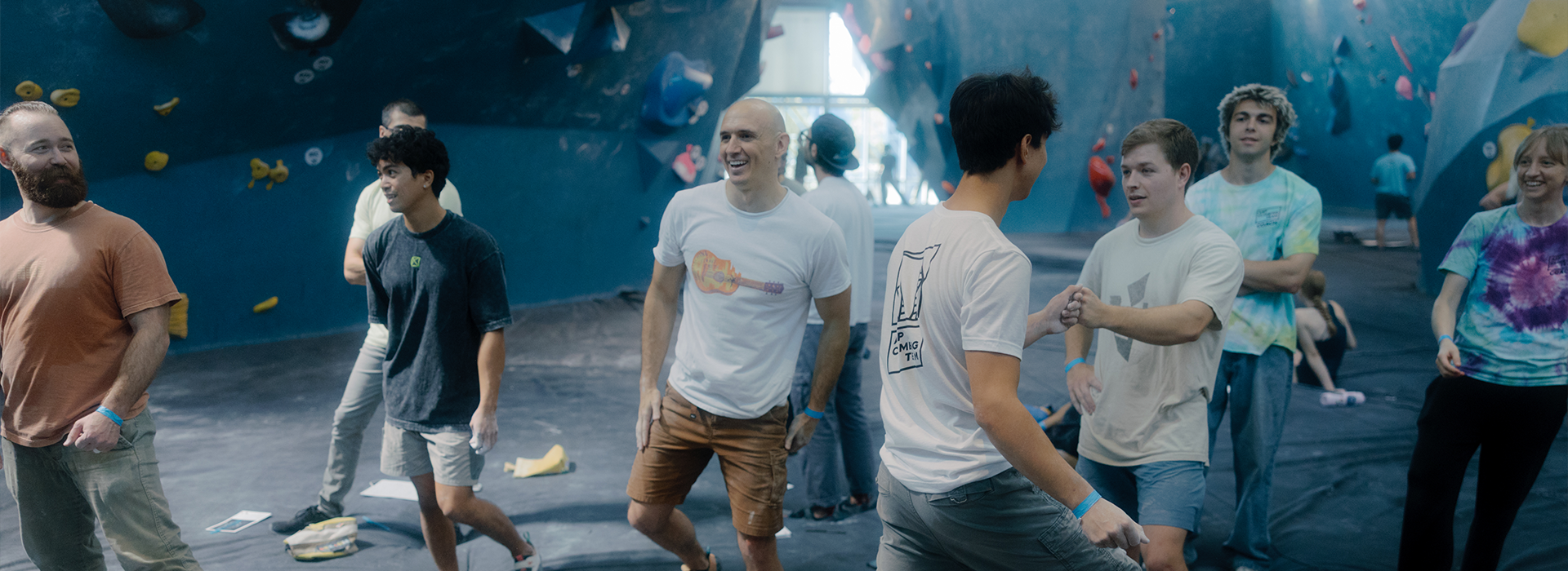 A group at Austin Bouldering Project socialize and prepare to climb in the vibrant gym. Some discuss group climbing reservations while others smile and explore the colorful holds and padded mats, ready for their next climb.