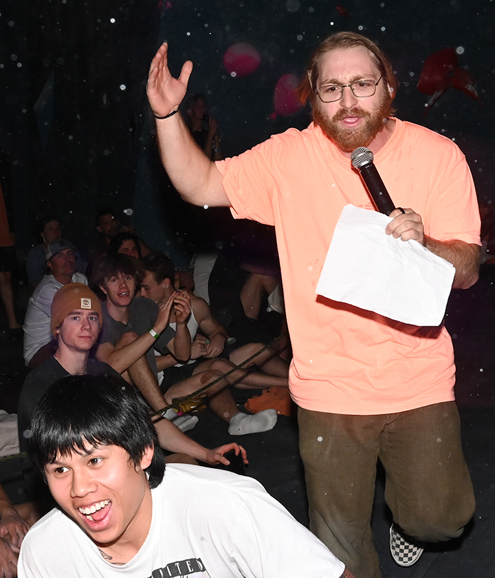 A man with a beard and glasses, wearing a bright orange shirt, holds a microphone and paper while speaking to an energetic Height Fight crowd in Austin. A smiling young man is in the foreground as the engaged audience listens closely.