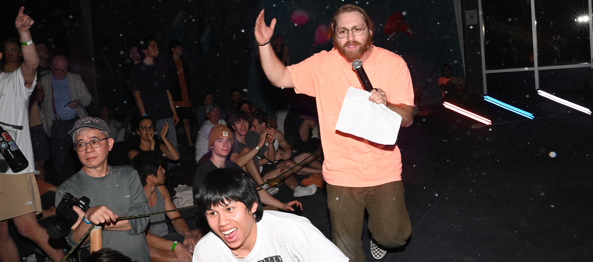 A bearded man in glasses holding a microphone and paper energetically addresses a crowd seated on the floor at Austins Height Fight, some smiling and interacting, in a dark indoor setting with colorful lights.