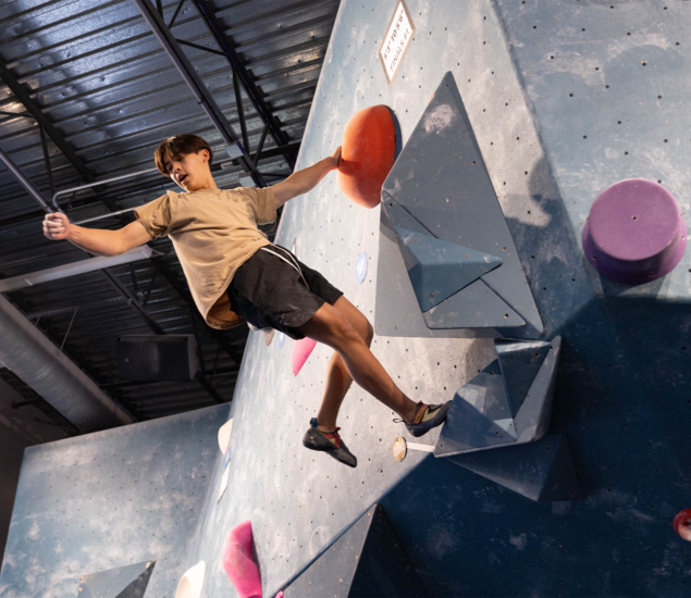 A person wearing a beige shirt and black shorts climbs an indoor bouldering wall in Austin, balancing on triangular holds with one foot off the wall under a metal ceiling with natural light—a true Height Fight challenge.