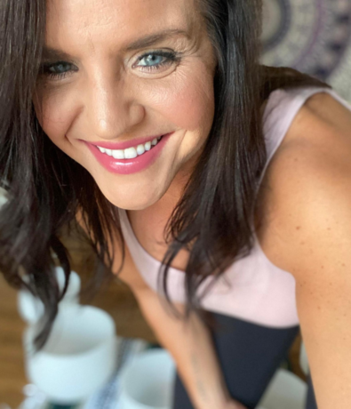 A woman with long dark hair smiles brightly at the camera. She is wearing a light pink sleeveless top and has clear blue eyes. The background is slightly blurred, showing part of a patterned tapestry and some white objects.