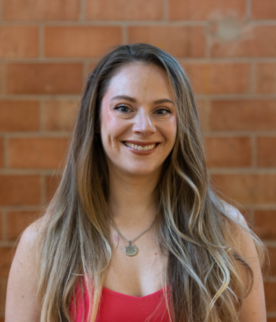 A woman with long, light brown hair and a necklace smiles at the camera. She is wearing a sleeveless, pink top and stands in front of a brick wall at an Austin climbing gym.