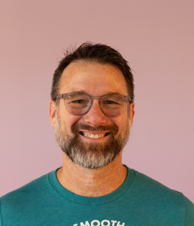 A smiling middle-aged man with glasses, short dark hair, and a salt-and-pepper beard, wearing a teal shirt, stands in front of a plain light purple background at an Austin Climbing Gym.