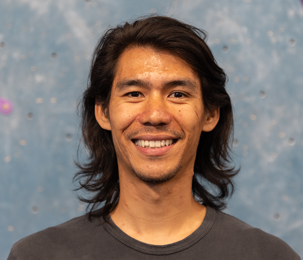 A man with long dark hair and a slight beard smiles at the camera. Wearing a dark shirt, he stands in front of a blue wall with small metal fixtures—reflecting his passion for Personal Climbing Coaching Austin.