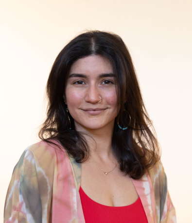 A person with long dark hair, wearing a nose ring, hoop earrings, a red top, and a tie-dye patterned outer garment, is smiling softly in front of a plain light background at an Austin climbing gym.
