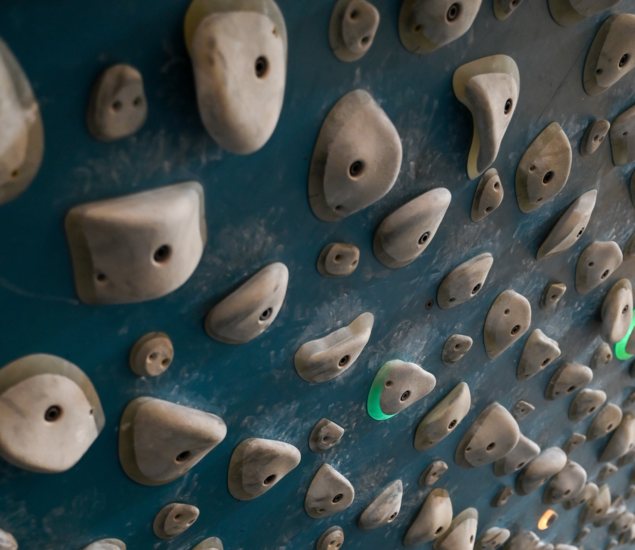 Close-up of a blue indoor climbing wall at Springdale, covered with various gray climbing holds and illuminated by green lights—perfectly arranged for bouldering or practicing routines on training boards.