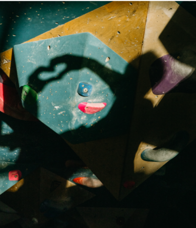 A shadow of a hand forming a heart shape is cast on a blue and yellow indoor climbing wall with colorful holds at an indoor rock climbing gym. Sunlight highlights the hand shadow against the geometric holds.