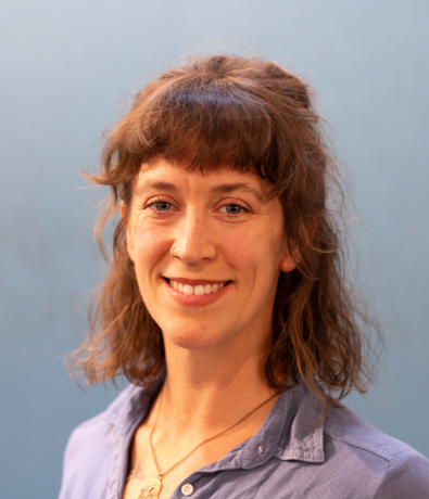 A woman with wavy brown hair and bangs smiles at the camera in an austin climbing gym. She is wearing a blue collared shirt and a necklace, with a soft blue background behind her.