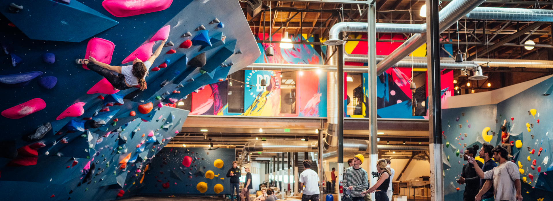 People climb colorful indoor bouldering walls in a lively, modern climbing gym with exposed ceilings, vibrant murals, and groups of people watching or preparing to climb.