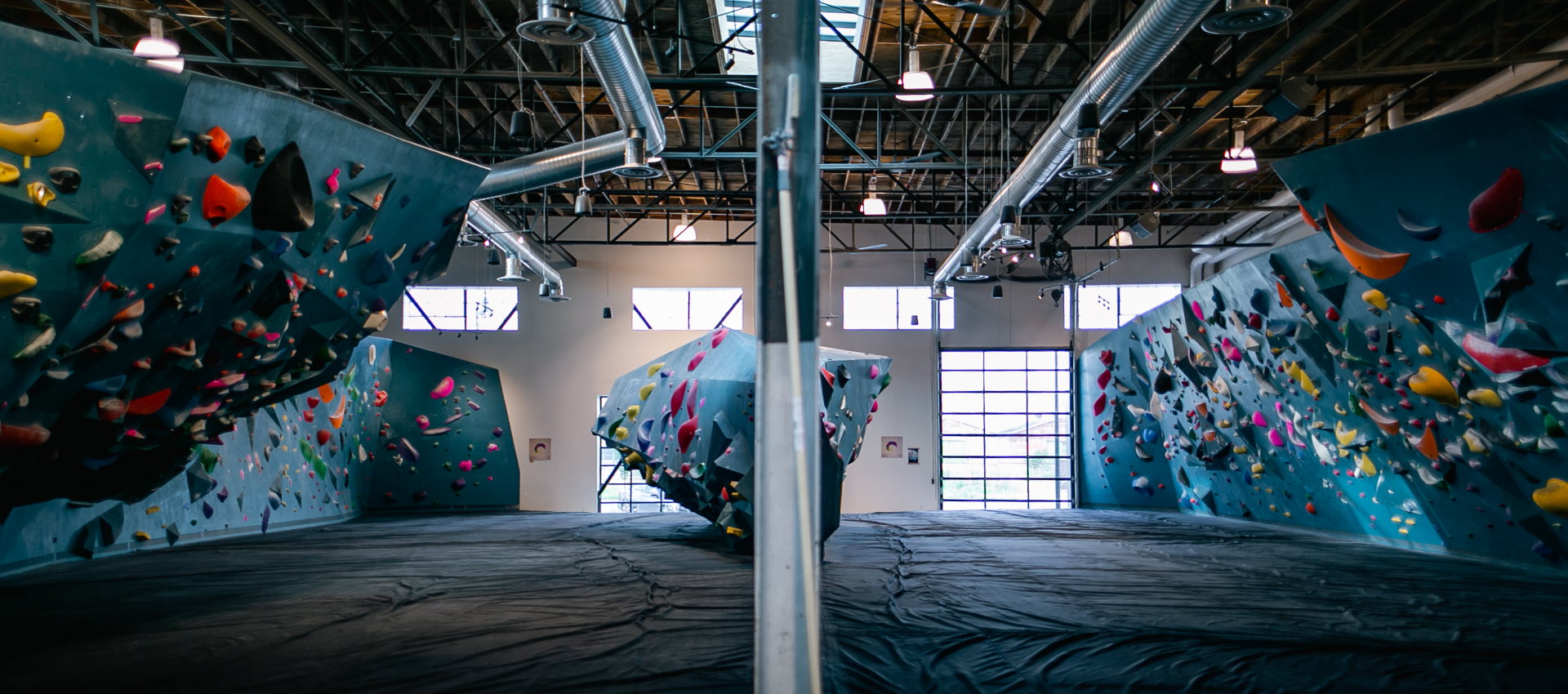 A modern indoor climbing gym with colorful climbing holds on angled blue walls, large windows letting in natural light, and exposed ceiling pipes. The scene is split by a reflective divider in the center.