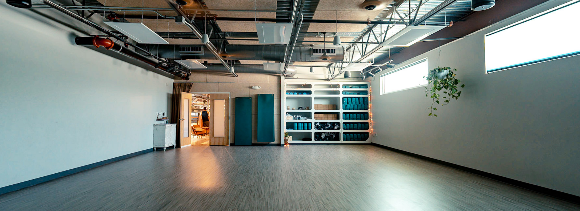A spacious, modern studio in an Austin climbing gym with wood flooring, shelves holding yoga mats and towels, a hanging plant, and exposed ceiling pipes. Natural light streams through two high windows; an open doorway leads to a warmly lit adjacent room.