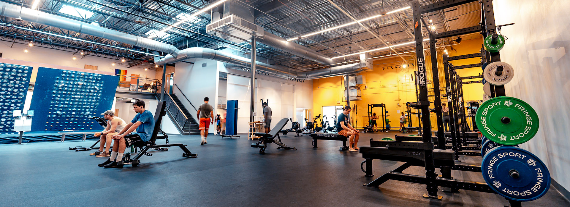 A spacious, modern gym with high ceilings, weightlifting racks, benches, and people working out. Wall-mounted equipment, weights, fitness machines, and features inspired by an Austin Climbing Gym fill the well-lit, open area.