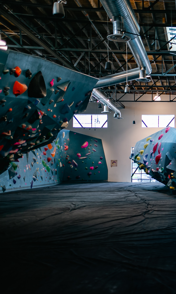 Indoor climbing gym with large bouldering walls covered in colorful holds, black padded flooring, exposed ceiling pipes, and natural light coming through windows in the background.