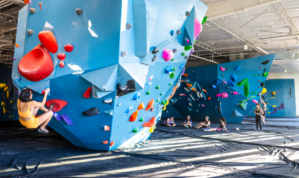 A person in a yellow outfit climbs a large blue indoor bouldering wall with colorful holds, while several people sit and watch in the spacious, brightly lit climbing gym.
