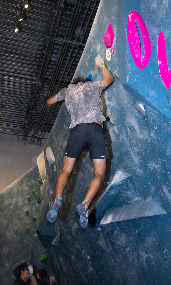A person wearing a gray shirt and black shorts climbs an indoor bouldering wall at a Minneapolis skills camp, using blue, pink, and white holds. The climber is in motion, reaching upward, while others watch from below.
