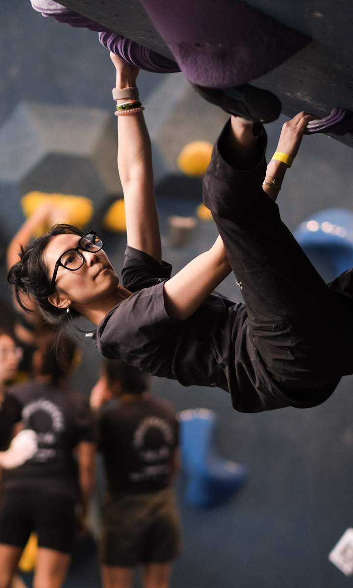 A woman with glasses and tied-back hair climbs an indoor bouldering wall, gripping a purple hold overhead, while people in the background observe a climbing lesson in progress.