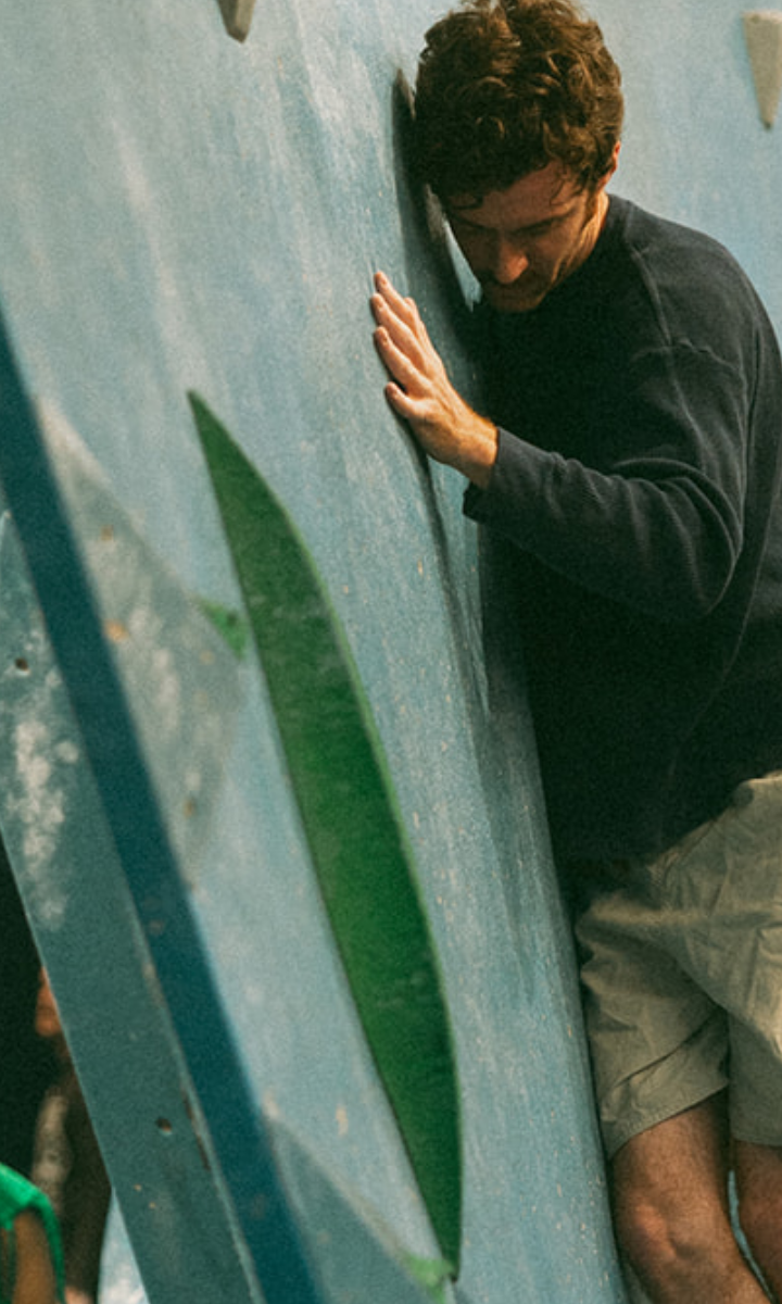 A man wearing a dark sweatshirt and light shorts enjoys an indoor climbing experience, pressing his body against the wall and using green holds for support.