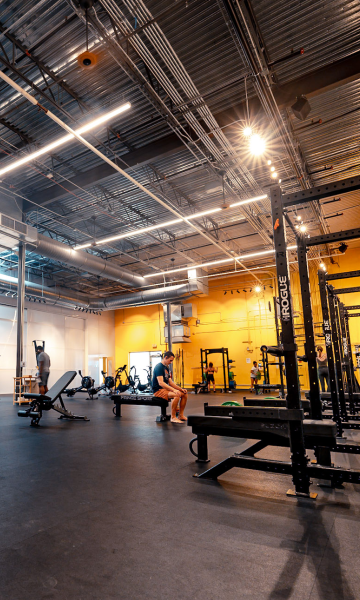 A spacious Austin climbing gym with high ceilings, yellow and white walls, exercise equipment, squat racks, and a few people working out. Bright overhead lights illuminate the open, modern workout area.