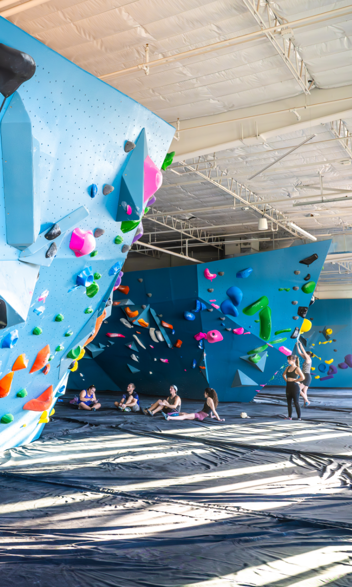 People are bouldering and sitting on mats in a bright indoor climbing gym with large blue climbing walls covered in colorful holds. Sunlight streams through the windows, casting shadows on the padded floor.
