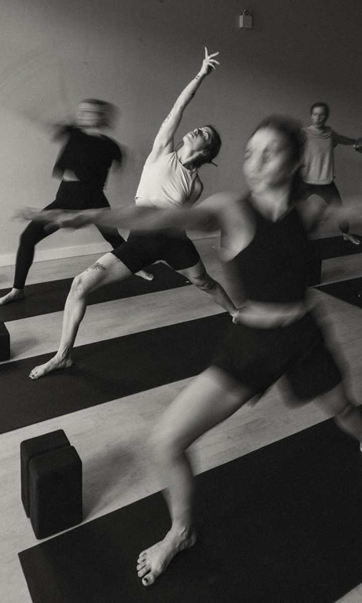 A group practices Vinyasa yoga indoors, captured in motion blur as they stretch on mats with yoga blocks nearby. The black and white photo, taken in a yoga studio, highlights dynamic movement and focus during yoga classes.