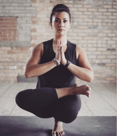 A woman practices yoga indoors, balancing on one leg in a seated pose with her hands pressed together at her chest. She wears black athletic clothing and bracelets, with a brick wall in the background.