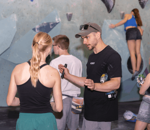 A man in a black shirt and cap discusses personal coaching with a woman in a tank top at an indoor climbing gym, while two others focus on the climbing wall nearby.
