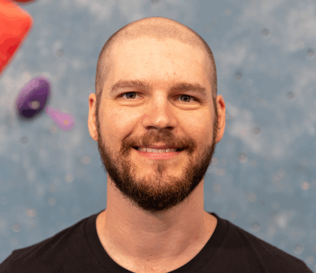 A bearded man with a shaved head smiles at the camera, standing in front of a blue indoor climbing wall with red and purple holds—ready to offer Personal Climbing Coaching in Austin.