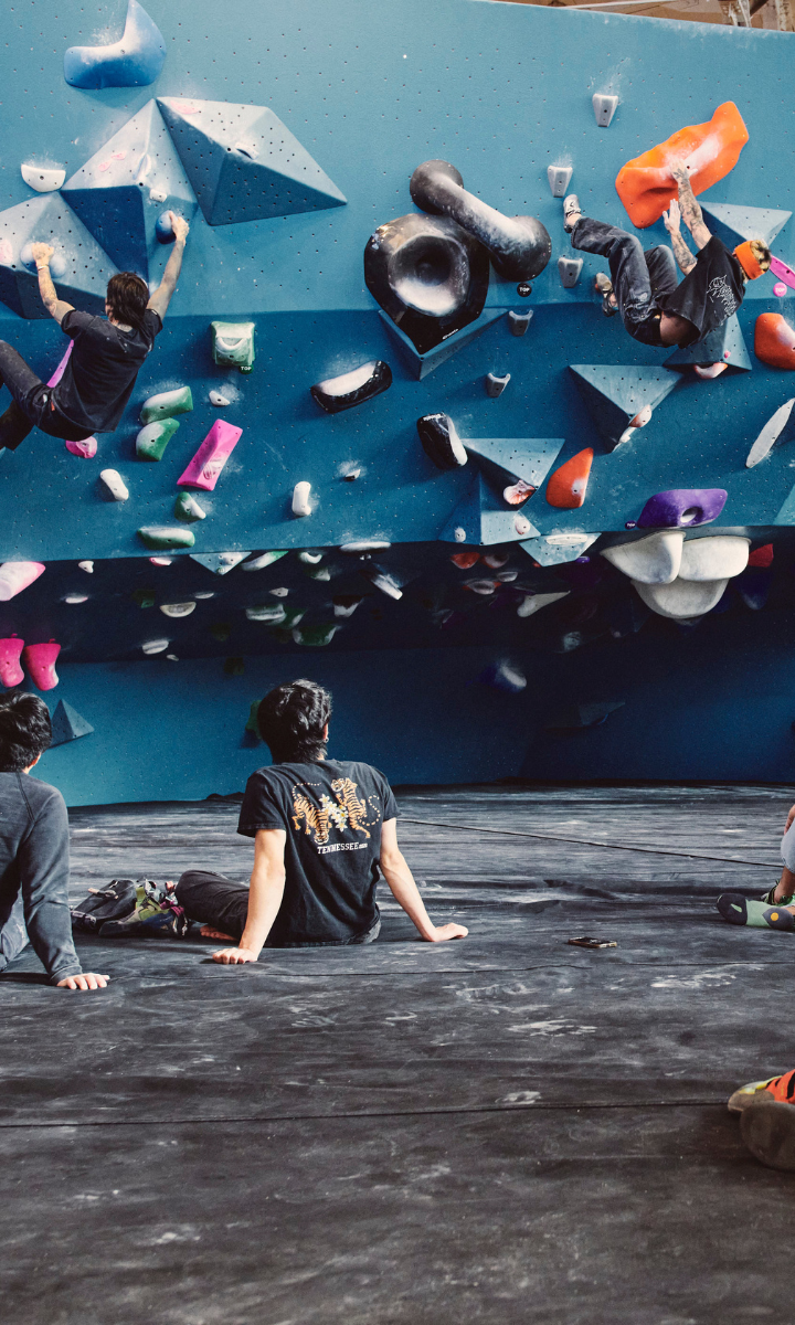 Several people are indoor bouldering on a climbing wall with colorful holds, while others sit on padded mats below, watching and resting—one of the exciting Brooklyn events happening today.