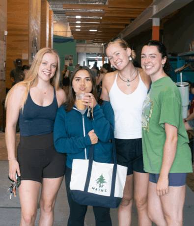 Four young women stand together indoors, smiling at the camera. One woman holds an iced coffee and a tote bag labeled MAINE. They are dressed casually in shorts and athletic wear. The background shows a busy, modern café.