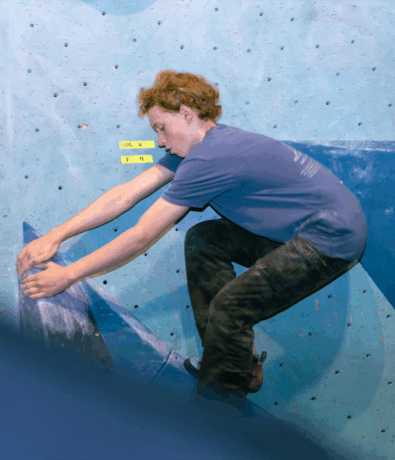 A person with short, curly red hair climbs an indoor blue bouldering wall, wearing a blue shirt and black pants. They are gripping large blue holds, with yellow route labels visible on the wall.