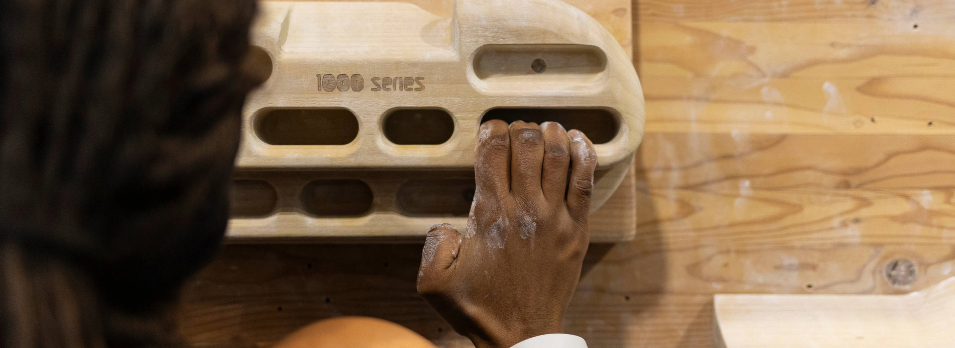 A person with chalked hands grips a wooden climbing hangboard mounted on a wooden wall in Minneapolis, practicing finger strength. The board is labeled 1000 Series—perfect for climbers seeking private coaching and serious training.