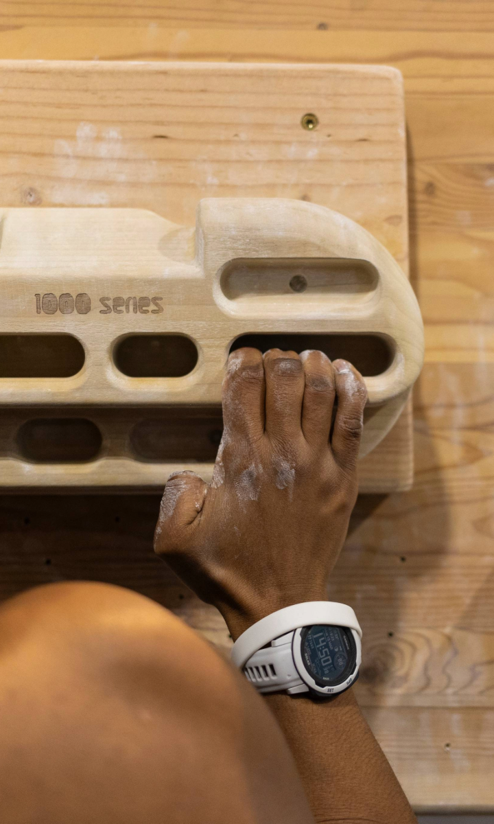 A person wearing a white sports watch grips a wooden hangboard with chalked fingers, practicing climbing holds on an indoor wooden wall—perfect training for those seeking Private Coaching in Minneapolis.