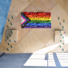 A climbing gym wall features a large climbing hold board painted with a rainbow pride flag and chevron, with green handholds on adjacent angled surfaces. Sunlight casts dramatic shadows across the floor, adding energy to the climbing space.