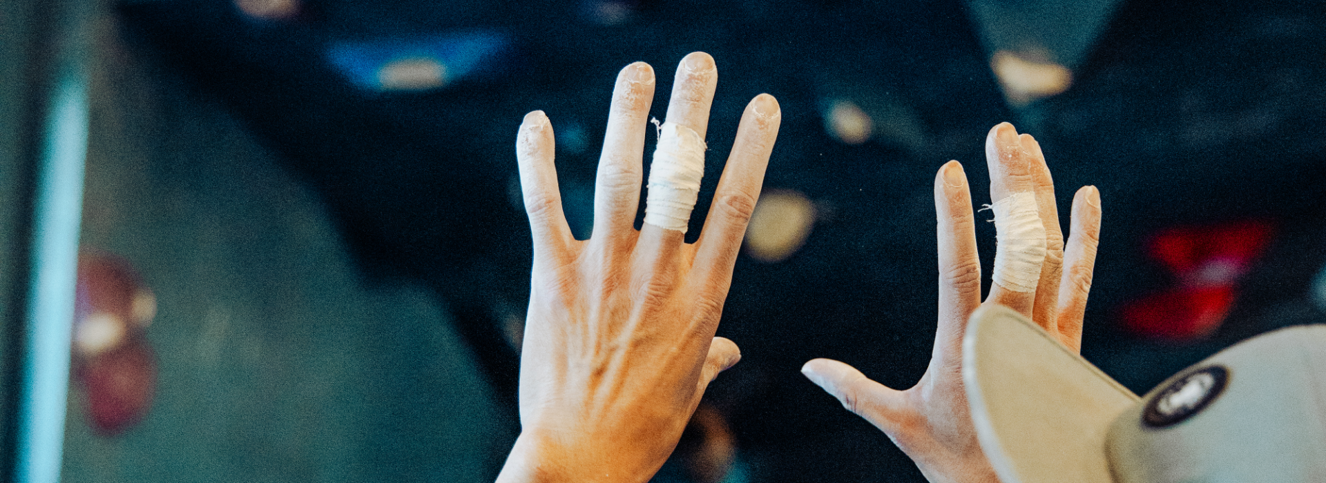 Two hands with fingers taped for support are raised against a blurred indoor rock climbing wall, suggesting preparation or recovery during Private Instruction. A person wearing a cap is partially visible in the foreground.
