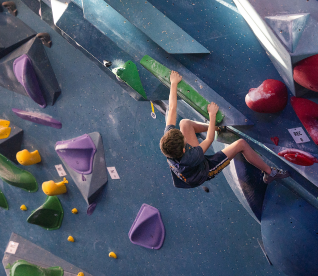 During school holidays in Brooklyn, a person wearing a blue shirt and shorts climbs an indoor bouldering wall with colorful holds, gripping a large green hold while reaching higher with one hand and supporting themselves with their legs.