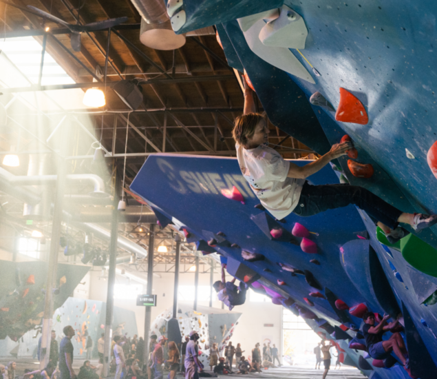 A person climbs a blue indoor bouldering wall with orange holds as others climb and watch in a spacious, sunlit climbing gym, gearing up for New Year 2025 promos available across all markets.