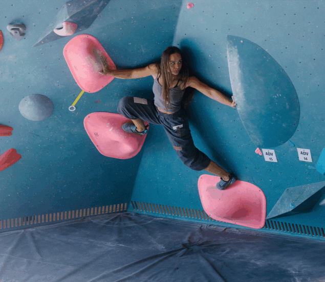 A woman in athletic clothing climbs an indoor bouldering wall, balancing on large pink holds with determination. Her focused expression suggests the benefits of personal coaching for climbers in all markets. The blue wall features various colorful holds.