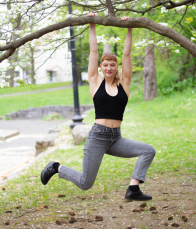 A person in a black tank top and gray jeans hangs from a tree branch with both hands, legs bent and smiling, in a park with green grass and trees in the background.