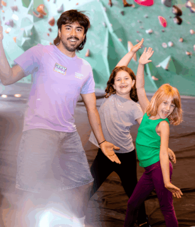 An adult and two young girls pose happily with playful, outstretched arms in front of an indoor climbing wall at an Austin climbing gym. The group smiles and appears energetic, enjoying their time together.