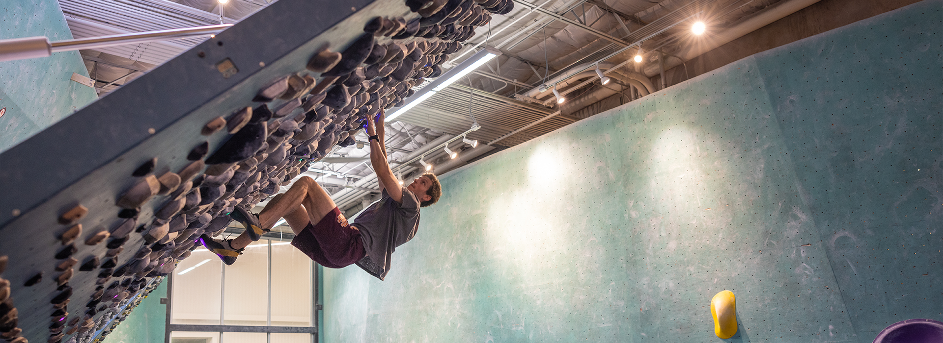 A person climbs an overhanging indoor bouldering wall at an Austin climbing gym, gripping holds with both hands and feet. The gym features high ceilings, overhead lights, and a green climbing wall in the background.